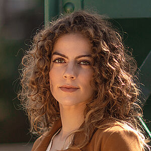 Portraitfoto von Alícia Navas Otero. Sie steht seitlich vor einer grünen Stahlstrebe. Ihr lockiges braunes Haar legt sich über ihre Schultern. Sie trägt einen camelfarbenen Blazer, darunter eine weiße Spitzenbluse.