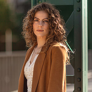 Portraitfoto von Alícia Navas Otero. Sie steht seitlich vor einer grünen Stahlstrebe. Ihr lockiges braunes Haar legt sich über ihre Schultern. Sie trägt einen camelfarbenen Blazer, darunter eine weiße Spitzenbluse. 