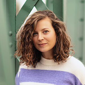 Portraitfoto von Julia Lindhorst-Apfelthaler. Sie hat schulterlanges welliges Haar. Sie trägt einen weiß-lilanen Pullover und lehnt an einer grünen Stahlstrebe.