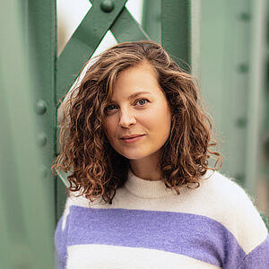 Portraitfoto von Julia Lindhorst-Apfelthaler. Sie hat schulterlanges welliges Haar. Sie trägt einen weiß-lilanen Pullover und lehnt an einer grünen Stahlstrebe.