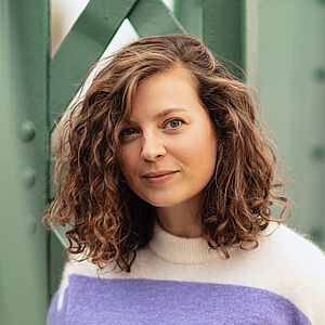 Portraitfoto von Julia Lindhorst-Apfelthaler. Sie hat schulterlanges welliges Haar. Sie trägt einen weiß-lilanen Pullover und lehnt an einer grünen Stahlstrebe.