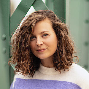 Portraitfoto von Julia Lindhorst-Apfelthaler. Sie hat schulterlanges welliges Haar. Sie trägt einen weiß-lilanen Pullover und lehnt an einer grünen Stahlstrebe.