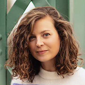 Portraitfoto von Julia Lindhorst-Apfelthaler. Sie hat schulterlanges welliges Haar. Sie trägt einen weiß-lilanen Pullover und lehnt an einer grünen Metallstrebe.