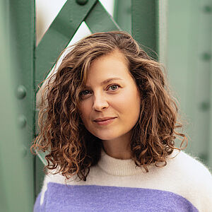 Portraitfoto von Julia Lindhorst-Apfelthaler. Sie hat schulterlanges welliges Haar. Sie trägt einen weiß-lilanen Pullover und lehnt an einer grünen Stahlstrebe.