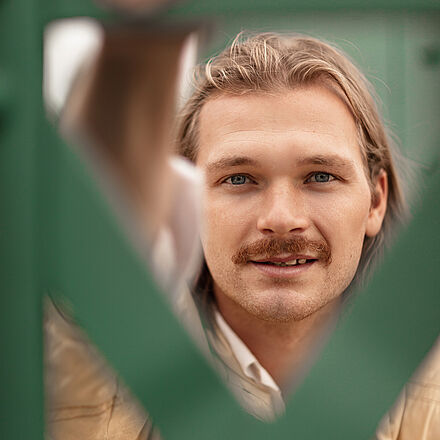 Portraitfoto von Leon Häder. Er blickt zwischen gekreuzten grünen Stahlstreben hindurch. Seine rechte Hand umfasst eine Strebe. Sein Mund ist leicht geöffnet. Er trägt langes blondes Haar, einen Schnurrbart und eine sandfarbene Jacke, darunter ein weißes Hemd.