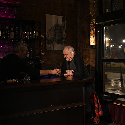 Der Mann sitzt an der Bar. Ihm wird ein Getränk gereicht.
