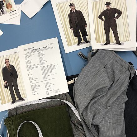 Auf einem Tisch liegen die Figurinen, eine Besetzungsliste, ein Maßband und Hosen.
