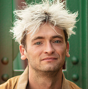 Portraitfoto von Philipp Haase. Er sitzt vor grünen Stahlstreben. Er trägt eine lederne beigefarbene Jacke, darunter ein weißes Hemd mit schwarzen Muster, das bis zur Brust aufgeknöpft ist. Sein Haar ist auf dem Kopf platinblond und steht empor.
