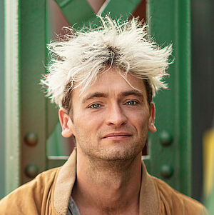 Portraitfoto von Philipp Haase. Er sitzt vor grünen Stahlstreben. Er trägt eine lederne beigefarbene Jacke, darunter ein weißes Hemd mit schwarzen Muster, das bis zur Brust aufgeknöpft ist. Sein Haar ist auf dem Kopf platinblond und steht empor.