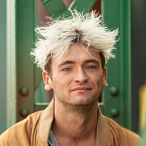 Portraitfoto von Philipp Haase. Er sitzt vor grünen Stahlstreben. Er trägt eine lederne beigefarbene Jacke, darunter ein weißes Hemd mit schwarzen Muster, das bis zur Brust aufgeknöpft ist. Sein Haar ist auf dem Kopf platinblond und steht empor.