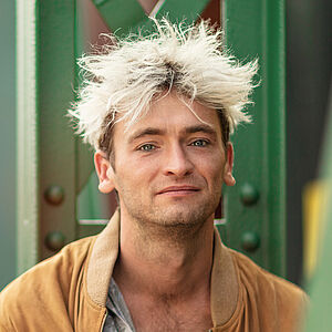 Portraitfoto von Philipp Haase. Er sitzt vor grünen Stahlstreben. Er trägt eine lederne beigefarbene Jacke, darunter ein weißes Hemd mit schwarzen Muster, das bis zur Brust aufgeknöpft ist. Sein Haar ist auf dem Kopf platinblond und steht empor.