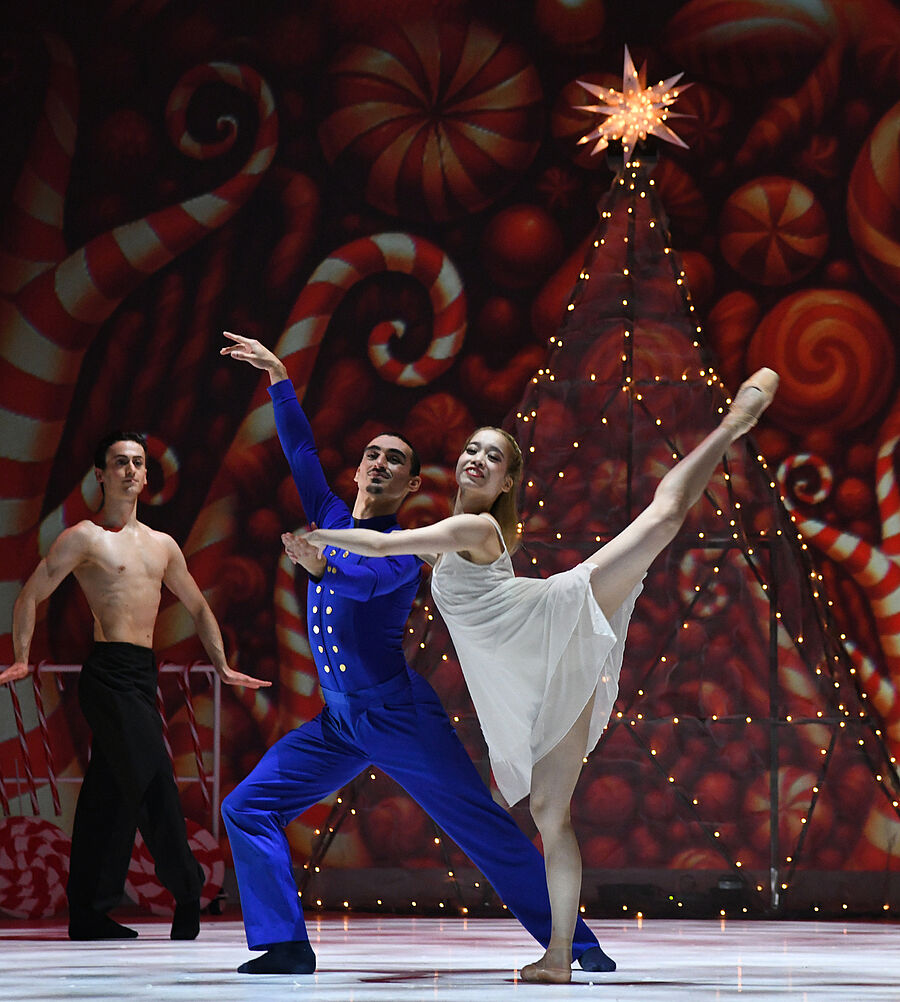 Im Hintergrund steht ein beleuchteter Tannenbaum, im Hintergrund sind Süßigkeiten zu sehen. Im Vordergrund stehen zwei Tänzer:innen und zwei Tänzer:innen stehen seitlich 
