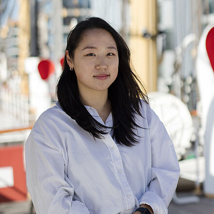 Portraitfoto von Chanmi Shin. Sie steht auf einem Schiff, das verschwommen erkennbar ist. Sie trägt eine weiße Bluse. Ihr schwarzes Haar liegt ihr auf den Schultern und wird vom Wind verweht. Sie lächelt leicht.