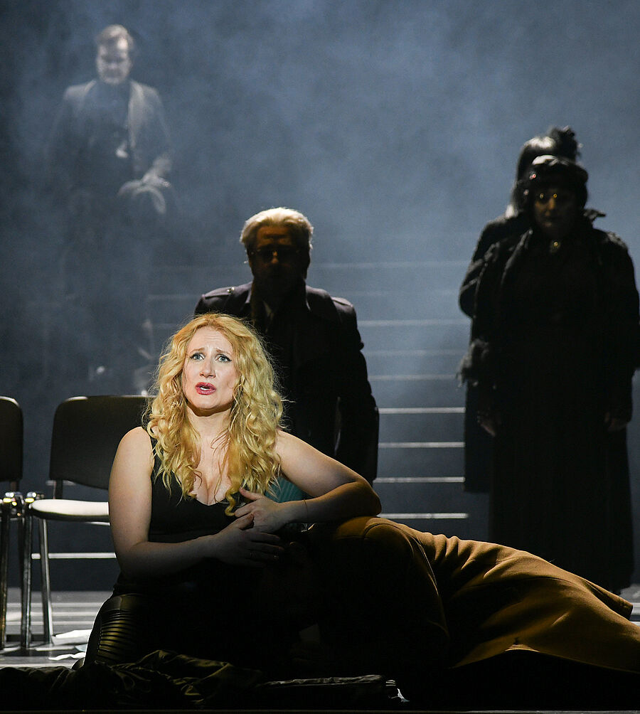 n der Mitte vom Bild liegt ein Mann im Schoß einer Frau. Im Hintergrund stehen mehrere Menschen im Schatten. 