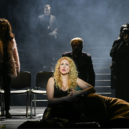 n der Mitte vom Bild liegt ein Mann im Schoß einer Frau. Im Hintergrund stehen mehrere Menschen im Schatten. 