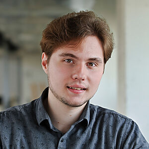 Portraitfoto von Anton Kononchenko. Er lächelt dezent. Er hat kurzes braunes Haar und Bart. Er trägt ein dunkelgraues Hemd mit schwarzen Aufdrucken. Der Hintergrund ist verschwommen.
