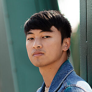 Portraitfoto von Ming-Hung Weng. Er steht seitlich und hat den Kopf zum Betrachter gerichtet. Er trägt sein dunkles Haar kurz sowie eine Jeansjacke. Hinter ihm eine grüne Stahlbrücke.