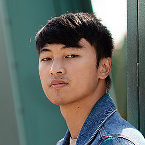 Portraitfoto von Ming-Hung Weng. Er steht seitlich und hat den Kopf zum Betrachter gerichtet. Er trägt sein dunkles Haar kurz sowie eine Jeansjacke. Hinter ihm eine grüne Stahlbrücke.