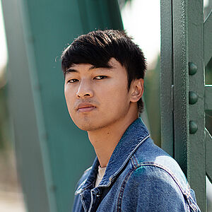 Portraitfoto von Ming-Hung Weng. Er steht seitlich und hat den Kopf zum Betrachter gerichtet. Er trägt sein dunkles Haar kurz sowie eine Jeansjacke. Hinter ihm eine grüne Stahlbrücke.