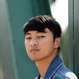 Portraitfoto von Ming-Hung Weng. Er steht seitlich und hat den Kopf zum Betrachter gerichtet. Er trägt sein dunkles Haar kurz sowie eine Jeansjacke. Hinter ihm eine grüne Metallbrücke.