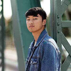 Portraitfoto von Ming-Hung Weng. Er steht seitlich und hat den Kopf zum Betrachter gerichtet. Er trägt sein dunkles Haar kurz sowie eine Jeansjacke. Hinter ihm eine grüne Stahlbrücke.