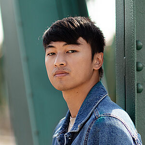 Portraitfoto von Ming-Hung Weng. Er steht seitlich und hat den Kopf zum Betrachter gerichtet. Er trägt sein dunkles Haar kurz sowie eine Jeansjacke. Hinter ihm eine grüne Stahlbrücke.