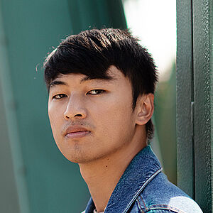 Portraitfoto von Ming-Hung Weng. Er steht seitlich und hat den Kopf zum Betrachter gerichtet. Er trägt sein dunkles Haar kurz sowie eine Jeansjacke. Hinter ihm eine grüne Metallbrücke.