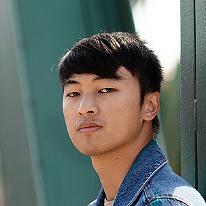 Portraitfoto von Ming-Hung Weng. Er steht seitlich und hat den Kopf zum Betrachter gerichtet. Er trägt sein dunkles Haar kurz sowie eine Jeansjacke. Hinter ihm eine grüne Metallbrücke.