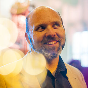 Portraitfoto von MacKenzie Gallinger. Er blickt schmunzelnd in die obere rechte Ecke. Er trägt einen hellbeigen Anzug, dazu ein dunkelblaues Hemd.