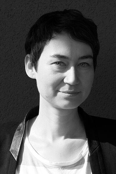 Schwarzweißes Portraitfoto von Judith Philipp. Sie blickt seitlich in die Kamera. Ihr Haar ist dunkel und kurz geschnitten. Sie lähelt dezent. Sie trägt ein weißes Oberteil, darüber eine dunkle Jacke.