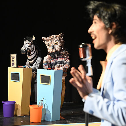 Eine Person steht am Mikro. Im Hintergrund stehen Personen am Pult.