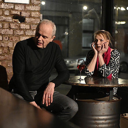 Eine Frau und ein Mann sitzen an einem Tisch. Sie trinken Wein. Er schaut weg, sie schaut ihn an.