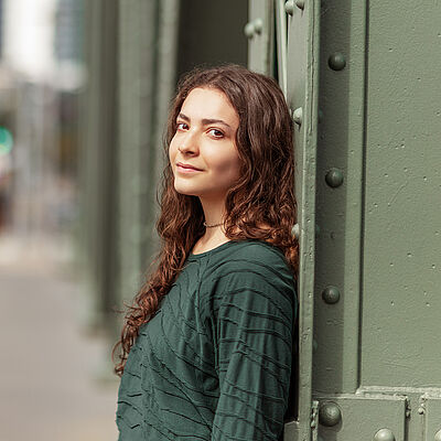 Portraitfoto von Helena Bröker. Sie lehnt an einer grünen Stahlstrebe. Ihre braunen Locken fallen ihr über die Schulter. Sie trägt einen dunkelgrünen Pullover.