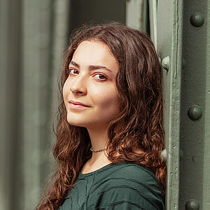 Portraitfoto von Helena Bröker. Sie lehnt an einer grünen Stahlstrebe. Ihre braunen Locken fallen ihr über die Schulter. Sie trägt einen dunkelgrünen Pullover.