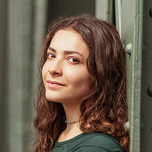 Portraitfoto von Helena Bröker. Sie lehnt an einer grünen Stahlstrebe. Ihre braunen Locken fallen ihr über die Schulter. Sie trägt einen dunkelgrünen Pullover.