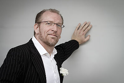 Portraitfoto von Jörg Seidel. Er ist in einen schwarzen Nadelstreifenanzug gekleidet. Dazu trägt er ein weißes Hemd. Sein hellgraues Haar ist zurückgegelt. Er trägt eine randlose Brille und grinst verschmitzt. Mit der linken Hand lehnt er an einer hellgrauen Wand.