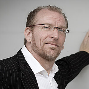 Portraitfoto von Jörg Seidel. Er ist in einen schwarzen Nadelstreifenanzug gekleidet. Dazu trägt er ein weißes Hemd. Sein hellgraues Haar ist zurückgegelt. Er trägt eine randlose Brille und grinst verschmitzt. Mit der linken Hand lehnt er an einer hellgrauen Wand. 