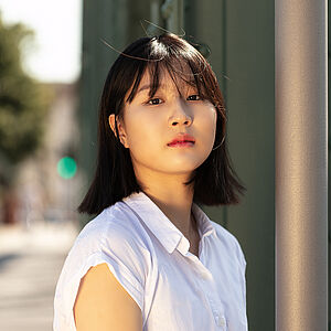 Portraitfoto von Miso Yun. Sie trägt ihr dunkles Haar offen. Es reicht bis zu den Schultern, ein Pony fällt ihr über die Stirn. Sie trägt eine weiße kurzärmlige Bluse und ist leicht seitlich gedreht. Neben ihr rankt eine graue Säule empor, im Hintergrund ist eine grüne Stahlbrücke zu erkennen. 
