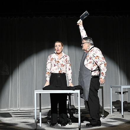 Eine Frau und ein Mann stehen an einem Tisch. Die Frau schaut nach vorne. Der Mann steht ihr zugewandt und hebt ein Messer in die Luft.