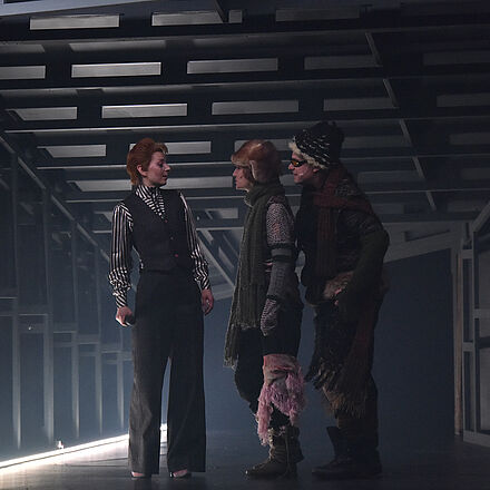 Drei Personen stehen im Mittelpunkt: links eine Frau, rechts eine Frau und ein Mann. Die beiden rechten Personen begutachten die linke Person.