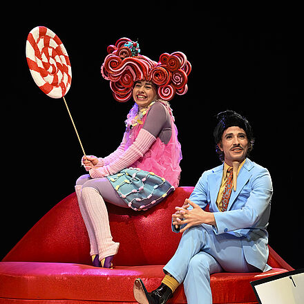 Zwei Personen sitzen auf einer lippenförmigen Bank. Die Person links trägt eine rote Perücke auf dem Kopf und hält einen großen Lutscher in der Hand. Die Person rechts trägt einen blauen Anzug.