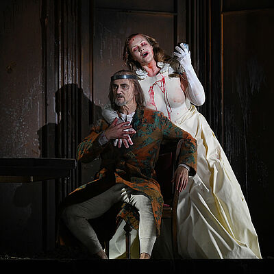 Lady Macbeth steht hinter Macbeth. Sie trägt ein weißes Kleid, an dem Blut herunterläuft. Er sitzt auf einem Stuhl am Tisch und trägt einen Morgenmantel sowie eine Krone. Ihr rechter Arm ist um Macbeth gelegt, er hält ihre Hand.