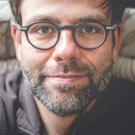 Portraitfoto von Toni Burkhardt. Er hat kurze dunkle Haare, einen Bart und trägt eine schwarze runde Brille.