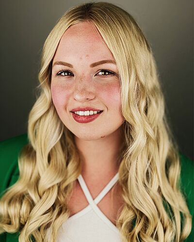 Portraitfoto von Marlene Mesa. Sie hat langes gewelltes blondes Haar, welches sie offen trägt. Sie lächelt mit geöffnetem rotem Mund. Sie trägt ein weißes Neckholder-Top, darüber einen grünen Cardigan.