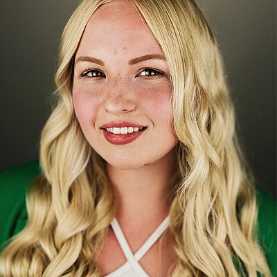 Portraitfoto von Marlene Mesa. Sie hat langes gewelltes blondes Haar, welches sie offen trägt. Sie lächelt mit geöffnetem rotem Mund. Sie trägt ein weißes Neckholder-Top, darüber einen grünen Cardigan.