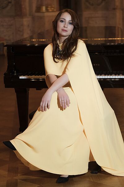 Portraitfoto von Regina Chernychko. Sie sitzt mit überschlagenen Beinen auf einem Klavierhocker. Hinter ihr steht ein schwarzer Flügel. Sie trägt ein hellgelbes Kleid. Sie trägt ihr dunkelbraunes Haar offen.