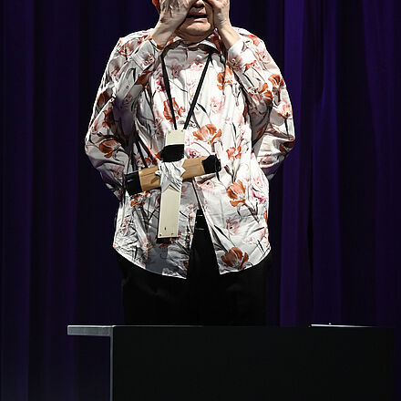 Im Mittelpunkt steht ein Mann mit einem Kreuz um den Hals. Unter seinen Achseln kommen zwei Hände hervor; sie liegen auf seinem Gesicht.