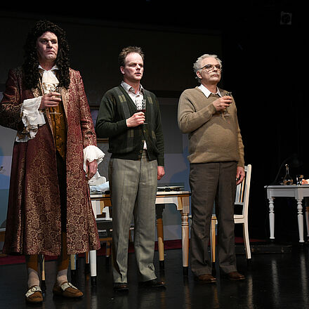 Im Hintergrund stehen zwei Tische. Am rechten Tisch sitzt eine Frau. Im Vordergrund stehen drei Männer, alle mit einem Weinglas in der Hand. Der linke Mann trägt Kleidung aus der Barock-Zeit.