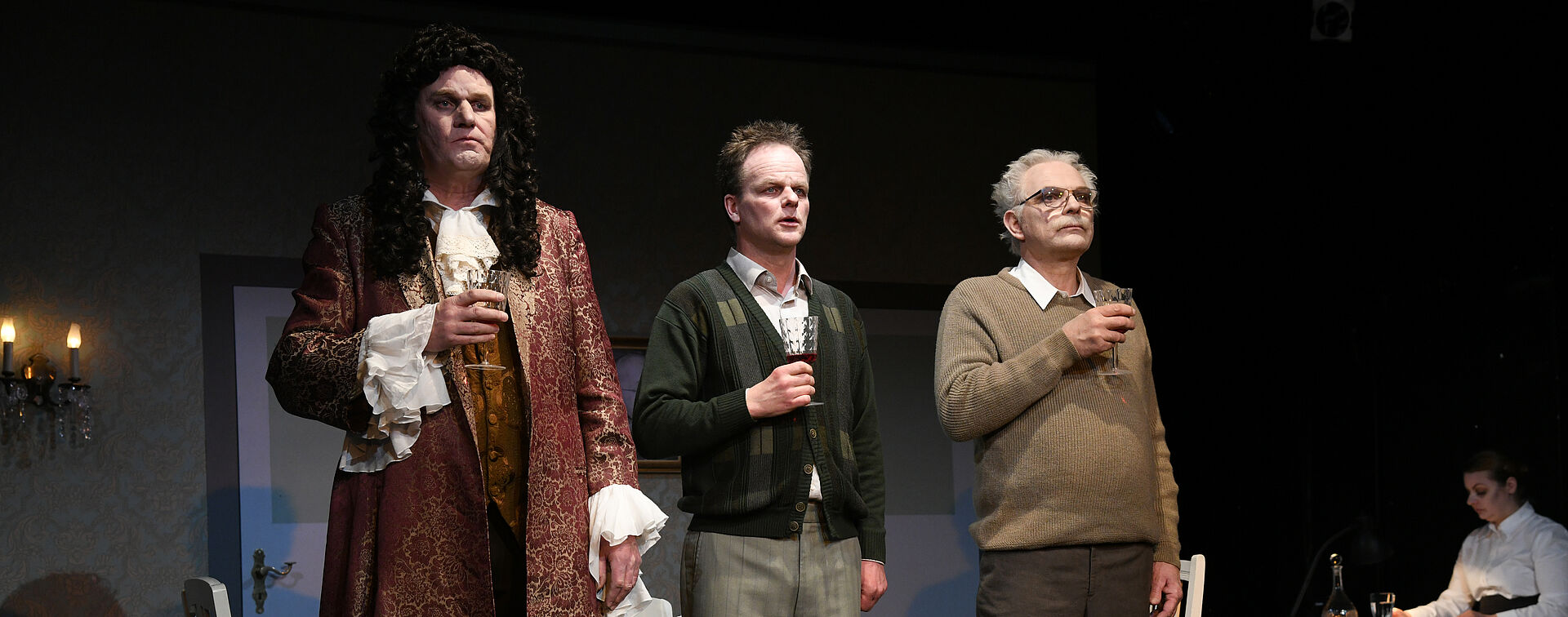Im Hintergrund stehen zwei Tische. Am rechten Tisch sitzt eine Frau. Im Vordergrund stehen drei Männer, alle mit einem Weinglas in der Hand. Der linke Mann trägt Kleidung aus der Barock-Zeit.