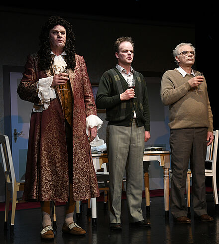 Im Hintergrund stehen zwei Tische. Am rechten Tisch sitzt eine Frau. Im Vordergrund stehen drei Männer, alle mit einem Weinglas in der Hand. Der linke Mann trägt Kleidung aus der Barock-Zeit.